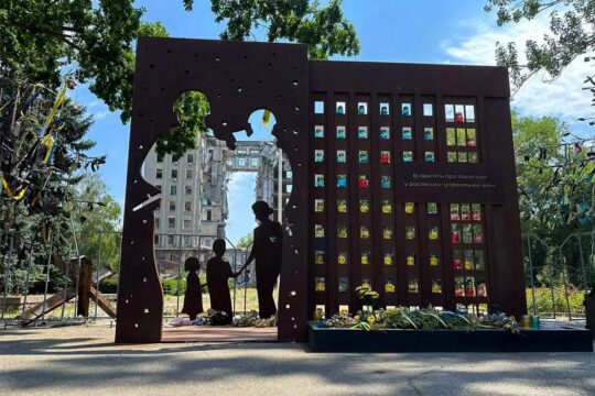 Monuments en mémoire des crimes de guerre en Ukraine. Photo : un monumemt dédié aux victimes de crimes de guerre représente une famille disparue qu'on voit au travers d'un mur percé. En arrière-plan, un bâtiment de plusieurs étages détruit.