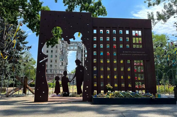 Monuments en mémoire des crimes de guerre en Ukraine. Photo : un monumemt dédié aux victimes de crimes de guerre représente une famille disparue qu'on voit au travers d'un mur percé. En arrière-plan, un bâtiment de plusieurs étages détruit.