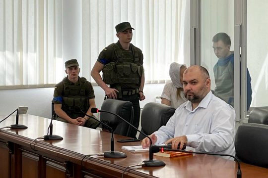 A lawyer (Viktor Ovsyannikov ) in a suit sits in the foreground. In the background are military personnel and a Vadim Shishimarin in the dock.