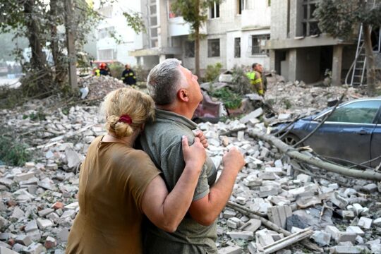 Dommages liés à la guerre en Ukraine : quelles réparations pour les victimes ? Photo : deux personnes enlacées regardent en l'air, au pied d'un immeuble d'habitation détruit par un missile russe.