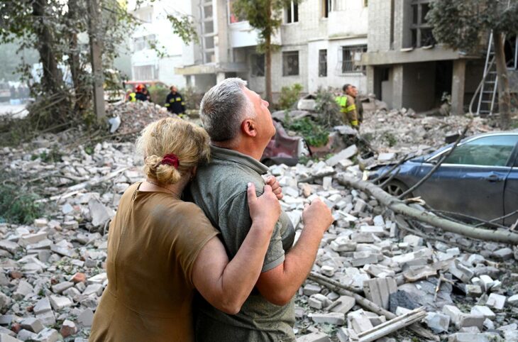Dommages liés à la guerre en Ukraine : quelles réparations pour les victimes ? Photo : deux personnes enlacées regardent en l'air, au pied d'un immeuble d'habitation détruit par un missile russe.