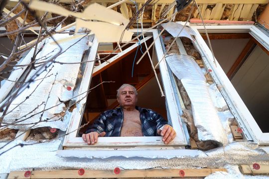 A man stands at the window of his partially destroyed house