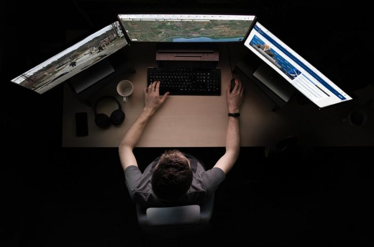 A man sitting in front of a desk works on 3 screens (satellite map + war image + ICC website)