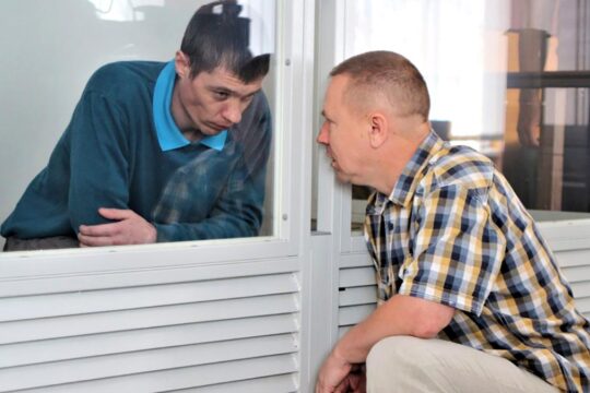 In Ukraine, who are these lawyers who defend the Russian occupiers when they are accused of crimes? Photo: A crouching man talks to another man (a soldier from Russia) through the glass of the dock.