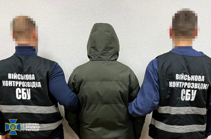 Un jeune agent recruté par la Russie (de dos, capuche sur la tête) est tenu de chaque côté par des policiers ukrainiens (de dos et floutés), face à un mur.