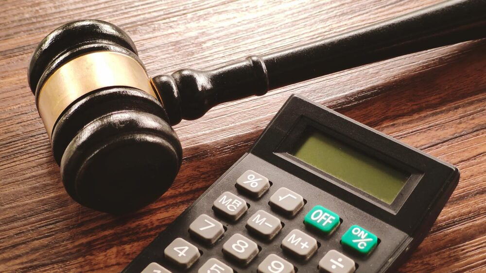 En cas d’acquittement, les équipes de défense et Orrön Energy, anciennement Lundin Petroleum, réclameront à l’État suédois une somme sans précédent dans l’histoire judiciaire suédoise. Photo : © Shutterstock Procès Lundin : la probabilité d'un acquittement de Ian Lundin et/ou Alexandre Schneiter laisse envisager un risque financier maximum. Photo : un marteau en bois (symbole de la justice) est posé sur une table, près d'une calculatrice.