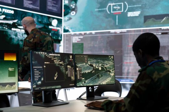 Military Artificial Intelligence (military AI) and international humanitarian law. Photo: Two soldiers seen from behind in a command room filled with screens showing naval targets viewed from the air.