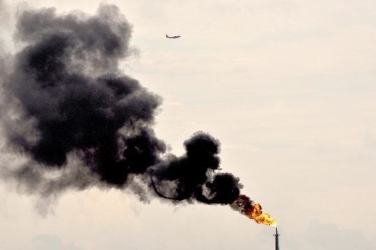 Climate change - Thick black smoke billows from an oil refinery. A plane flies in the sky.