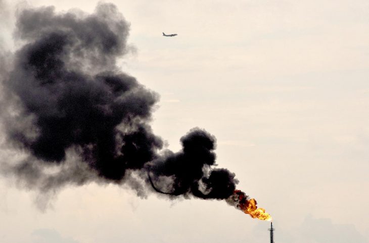 Climate change - Thick black smoke billows from an oil refinery. A plane flies in the sky.