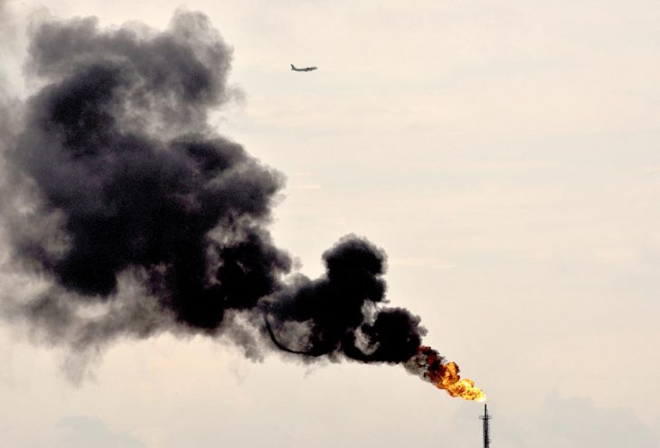 Climate change - Thick black smoke billows from an oil refinery. A plane flies in the sky.