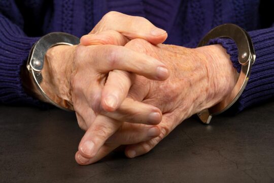 How does international justice deal with defendants accused of mass crimes when they are elderly? Photo: Hands clasped together of an elderly person wearing handcuffs